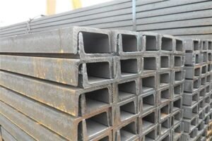 A stack of steel channel sections neatly arranged on the workshop floor, waiting to be cut by the bandsaw blade.